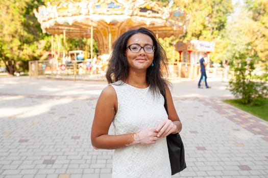 Happy African American woman with skin problem in eyeglasses standing at amusement park. Portrait of smiling lady with Vitiligo disease in white dress looking at camera.