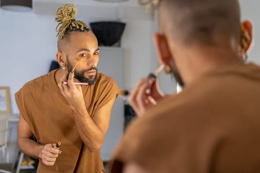 Black male gay applying make up looking mirror standing back to camera. Stylish homosexual man in dressing room doing make-up on face