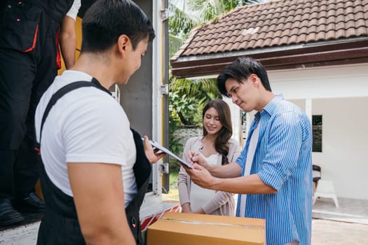 Professional movers help a satisfied couple sign the delivery checklist after furniture lifting. Employee teamwork assures customer contentment. Moving Day Concept