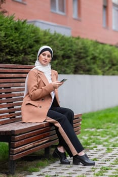 Young woman in hijab uses a smartphone while sitting on a bench outdoors