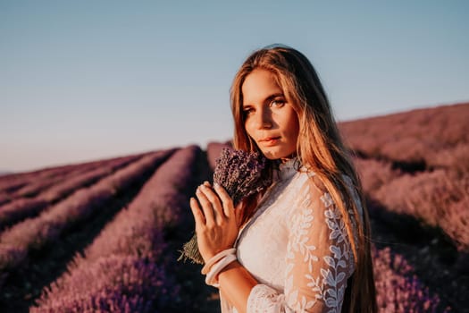 Close up portrait of young beautiful woman in a white dress and a hat is walking in the lavender field and smelling lavender bouquet.