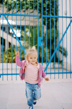 Little smiling girl runs from a metal gate along a paved path waving her arms. High quality photo