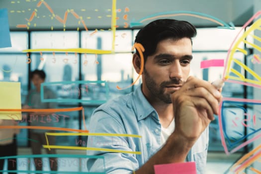 Portrait of young handsome businessman writing marketing idea by using colorful marker on glass wall with graph and mind map. Manager sharing idea or financial strategy. Business plan. Tracery.