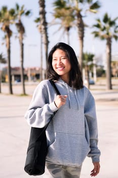 smiling young asian woman dressed in casual clothes walking with yoga mat hanging on her arm, sport and healthy lifestyle concept