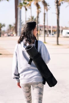 rear view of a smiling young asian woman dressed in casual clothes walking with yoga mat hanging on her arm, sport and healthy lifestyle concept