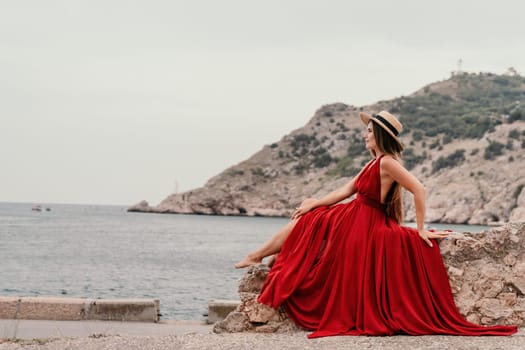 Side view a Young beautiful sensual woman in a red long dress posing on a volcanic rock high above the sea during sunset. Girl on the nature on overcast sky background. Fashion photo