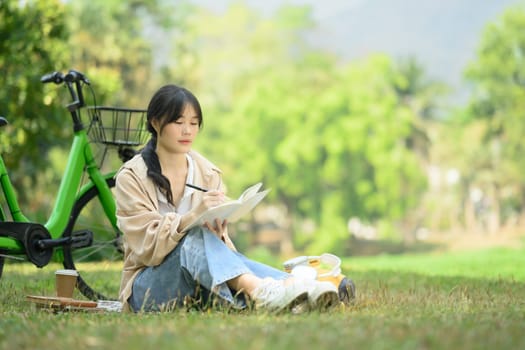 Full length of concentrated Asian female student reading book on campus lawn next to her bicycle.