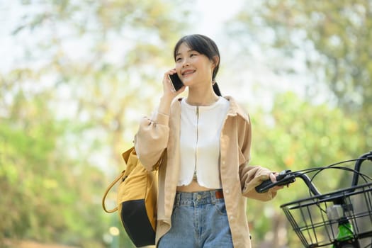 Positive young woman in casual clothes talking on mobile phone and standing near bicycle.