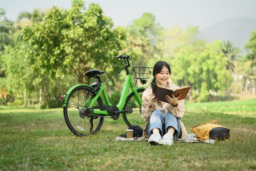 Peaceful young woman relaxing on green grass and reading book during summer day.