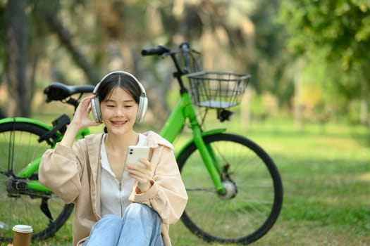 Smiling joyful young woman listening music over smartphone sitting on grass near her bicycle.