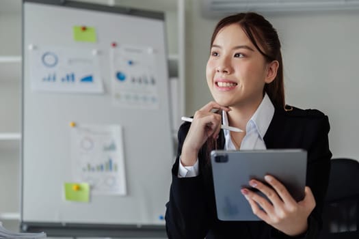 A woman in a business suit is sitting at a desk with a tablet and a pen. She is smiling and she is focused on her work. The scene suggests a professional and productive atmosphere