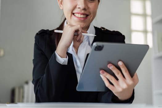A woman in a business suit is sitting at a desk with a tablet and a pen. She is smiling and she is focused on her work. The scene suggests a professional and productive atmosphere