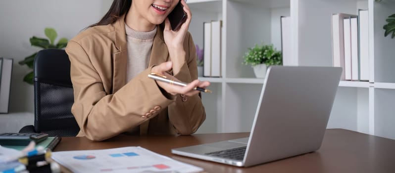 Young Asian business woman sits on the phone in an online business meeting using a laptop in a modern home office decorated with shady green plants..