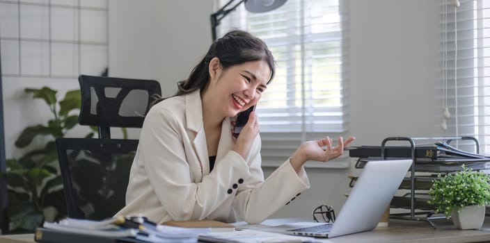 Young Asian business woman sits on the phone in an online business meeting using a laptop in a modern home office decorated with shady green plants..