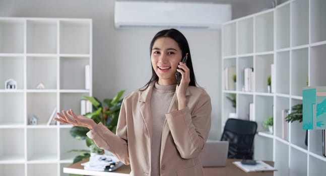 Young Asian business woman sits on the phone in an online business meeting using a laptop in a modern home office decorated with shady green plants..