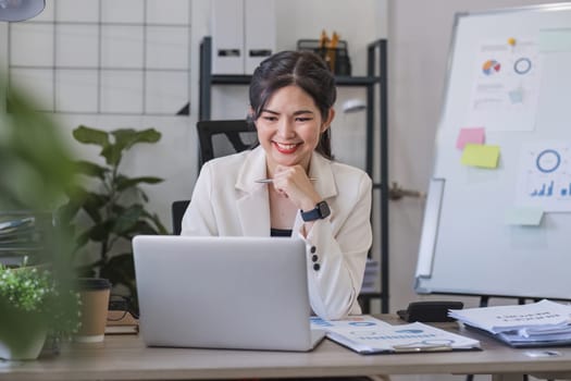 Asian businesswoman or accountant happily sitting and working with laptop on finance and business administration. On the desk in the office decorated with green plants..