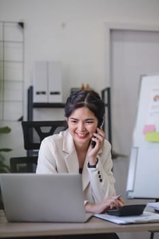 Young Asian business woman sits on the phone in an online business meeting using a laptop in a modern home office decorated with shady green plants..