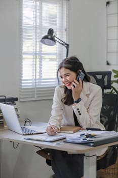 Young Asian business woman sits on the phone in an online business meeting using a laptop in a modern home office decorated with shady green plants..