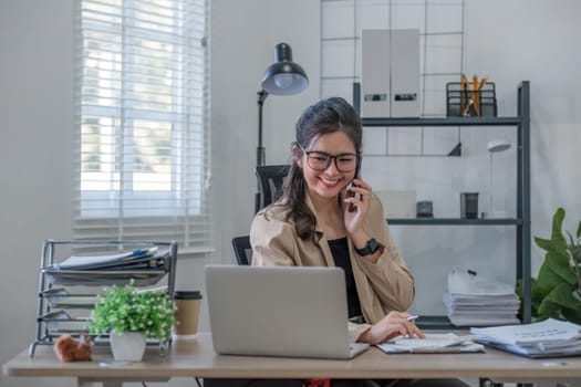 Young Asian business woman sits on the phone in an online business meeting using a laptop in a modern home office decorated with shady green plants..