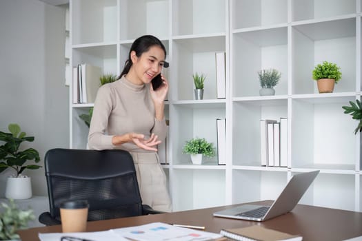 Young Asian business woman sits on the phone in an online business meeting using a laptop in a modern home office decorated with shady green plants..