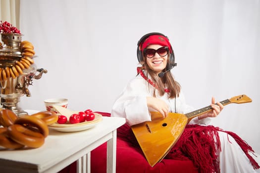 A girl in traditional national russian dress with a scarf and shawl, with musical balalaika and modern headphones with a microphone and glasses at a set table for Easter and Maslenitsa