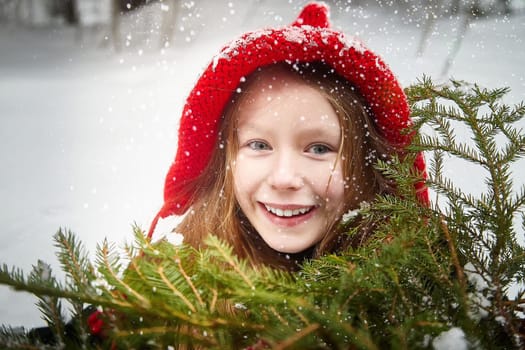 Cute little girl in red cap or hat and black coat with basket of green fir branches in forest and snow on winter day. Young girl went to gather firewood in cold. Fun and fairytale on photo shoot