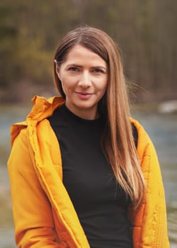 Portrait of young woman in yellow jacket, smiling, long hair down, blurred trees and river background