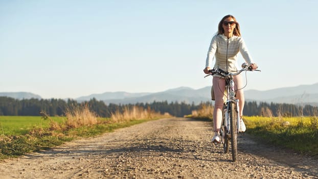 Young woman in jacket and sunglasses rides a bicycle over country road, afternoon sun shines on blurred background, space for text left side