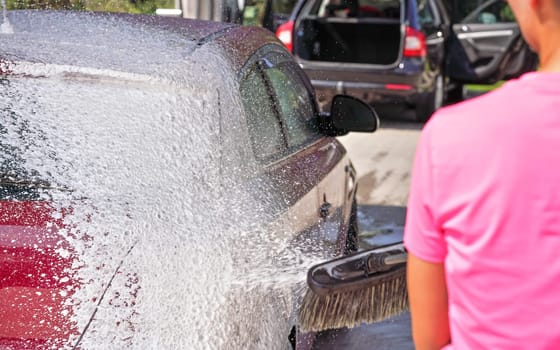 Red car washed in self serve carwash, view from rear of young woman spraying foam from high pressure hose, detail on small white soap drops flying