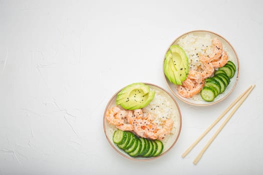 Two white ceramic bowls with rice, shrimps, avocado, vegetables and sesame seeds and wooden sticks on white concrete stone background top view. Healthy asian style poke bowl, space for text.
