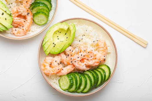 Two white ceramic bowls with rice, shrimps, avocado, vegetables and sesame seeds and wooden sticks on white concrete stone background top view. Healthy asian style poke bowl.