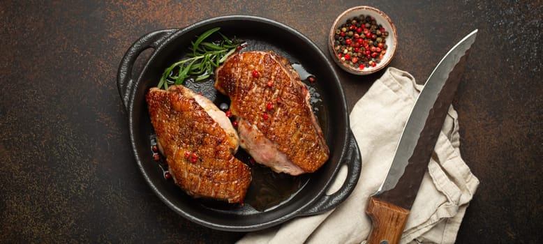 Two roasted duck breast fillets with crispy skin, with pepper and rosemary, top view in black cast iron pan with knife, dark brown concrete rustic background.