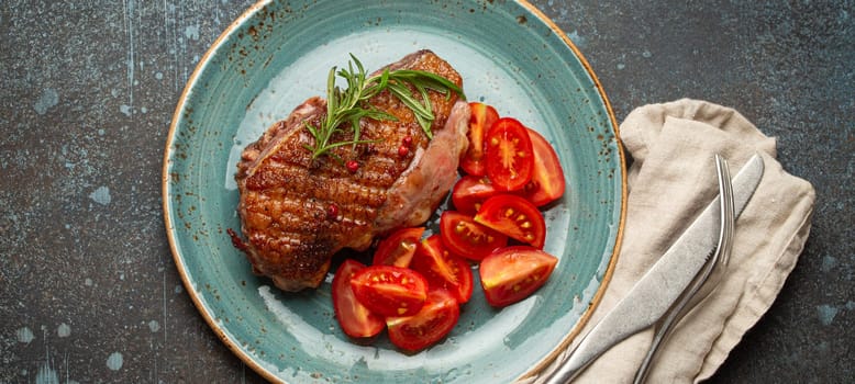 Delicious roasted duck breast fillet with golden crispy skin, with pepper and rosemary, top view on ceramic blue plate served with cherry tomatoes salad, rustic concrete rustic background