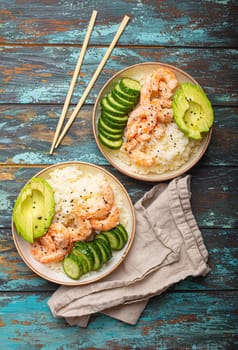 Two white ceramic bowls with rice, shrimps, avocado, vegetables and sesame seeds and chopsticks on colourful rustic wooden background top view. Healthy asian style poke bowl.