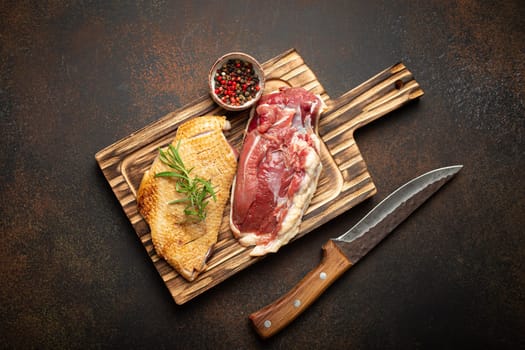 Two raw uncooked duck breast fillets with skin, seasoned with salt, pepper, rosemary top view on wooden cutting board with knife, dark brown concrete rustic background.