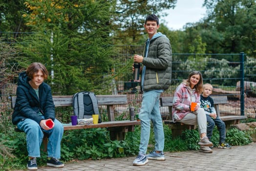 Happy Family drinking tea from a thermos in the forest. Photo of cute charming mother with childrens dressed casual outfit walking sitting bench drinking tea smiling outside city park