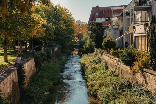 Beautiful small river with clean and clear water front of colorful autumn trees and small old town on the hill agaist nice blue and clouds sky during autumn in Europe.