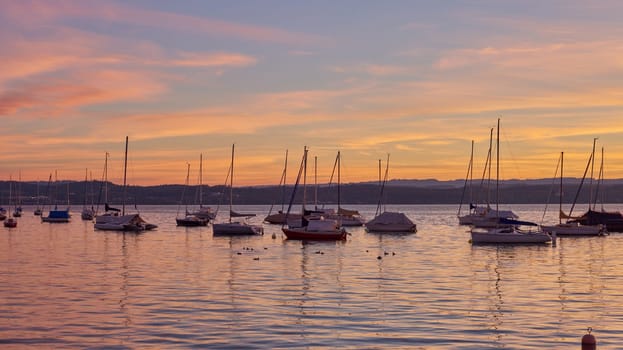 Bodensee Lake Sunrise Panorama. Morning Sunlight Over Tranquil Waters. Witness the mesmerizing dawn over Germany's Bodensee Lake, captured from a boat dock. Embrace the tranquil beauty of the early morning as the sun rises, casting a soft glow on the landscape. The peaceful scene features boats, yachts, and a charming water shack set against a backdrop of a captivating sky. Clouds delicately reflect on the calm water, creating a serene atmosphere. Immerse yourself in the serene beauty of a lakeside sunrise. Explore the harmony of nature, technology, and production as the day unfolds by the lake.