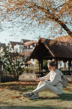 Young fashionable teenage girl with smartphone in park in autumn sitting at smiling. Trendy young woman in fall in park texting. Retouched, vibrant colors. Beautiful blonde teenage girl wearing casual modern autumn outfit sitting in park in autumn. Retouched, vibrant colors, brownish tones.