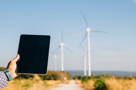 Technician tablet in hand at windmill farm. Engineer ensures clean energy. Expertise in turbine efficiency and global electricity control portrayed confidently.