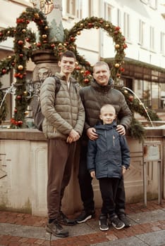 Joyful Family Portrait: Father and Two Sons by Festive Vintage Fountain. Capture the essence of familial happiness with this heartwarming image featuring a handsome father with his two sons standing against the backdrop of a festively decorated vintage fountain. The image beautifully blends the joy of the holiday season with the timeless charm of family togetherness. The father is 44 years old, the elder son is 17, and the younger one is 8, creating a memorable and multigenerational festive scene.