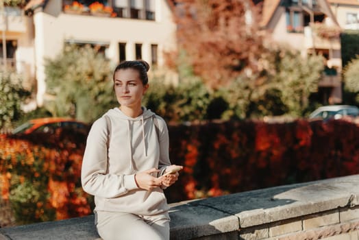 Young fashionable teenage girl with smartphone in park in autumn sitting at smiling. Trendy young woman in fall in park texting. Retouched, vibrant colors. Beautiful blonde teenage girl wearing casual modern autumn outfit sitting in park in autumn. Retouched, vibrant colors, brownish tones.