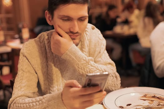 Portrait of one young handsome caucasian brunette man in a knitted sweater with a phone in his hands looking at the screen and sitting at a table in a restaurant, close-up side view.Men lifestyle concept,using technology.