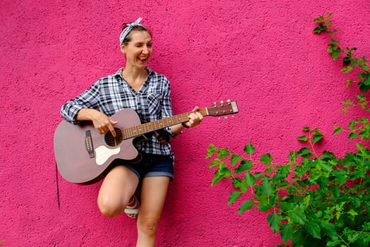 Young woman playing acoustic guitar on pink wall and green leaves background. Hipster girl portrait on street. Positive girl with guitar download