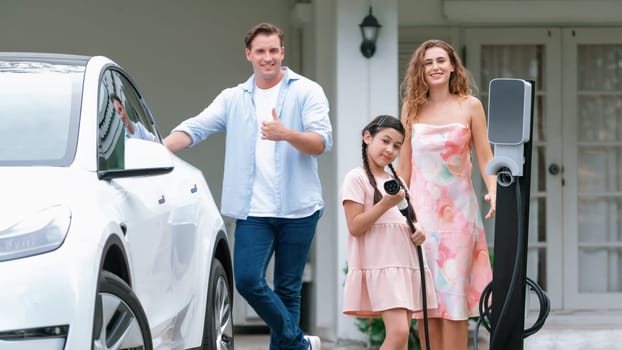 Modern family with environmental conscious recharging EV car with home EV charging station, Happy family with little young girl holding and pointing EV charger at camera. Synchronos