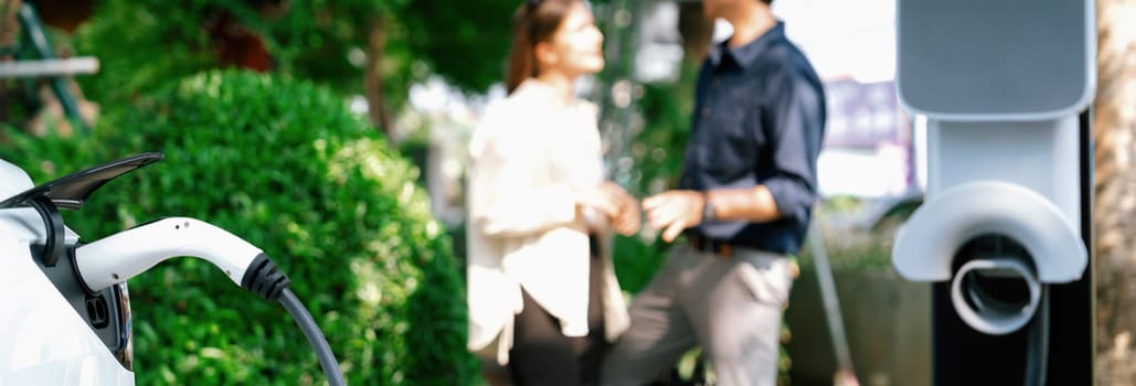 Young couple recharge electric car battery from charging station in green city park in springtime. Rechargeable EV car for sustainable environmental friendly urban travel lifestyle. Panorama Expedient