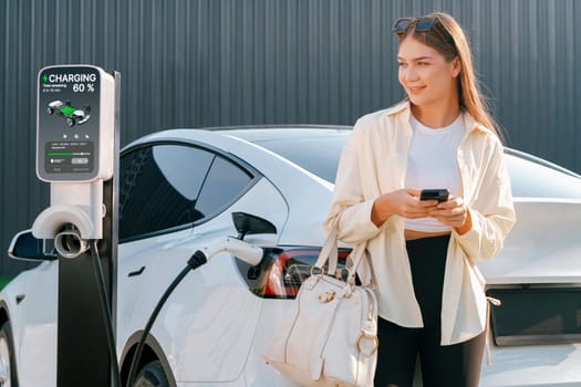 Young woman holding shopping bag and use smartphone to pay for electricity for recharging EV car battery from charging station at city mall parking lot. Modern woman go shopping by eco car. Expedient