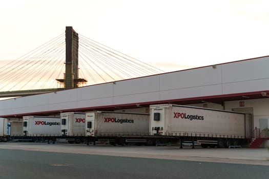 Seville, Spain - June 1, 2023: Several XPO Logistics trailers parked at a loading dock with a cable-stayed bridge in the background, captured at dusk.