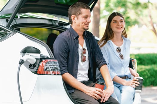 Lovely young couple drinking coffee while recharging battery for electric car during road trip travel EV car in natural forest or national park. Eco friendly travel during vacation and holiday. Exalt