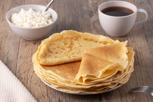 A stack of thin pancakes, a plate with cottage cheese and coffee on a wooden table.